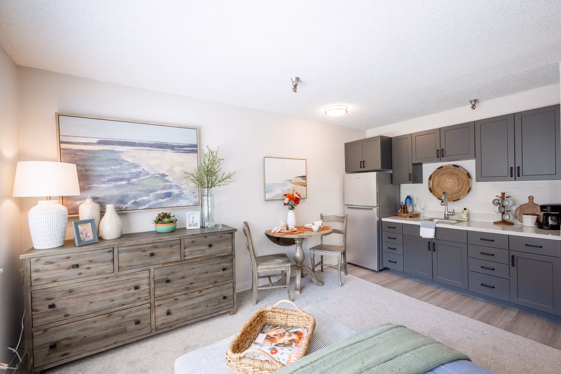 Well-lit studio interior showing a kitchenette with gray cabinets, a small dining table, wooden dresser, lamp, and wall art.