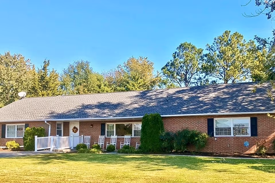 Single-story brick building with a gray shingled roof, white door with a wreath, several white rocking chairs on a porch with a white railing, surrounded by green grass and trees under a clear blue sky.