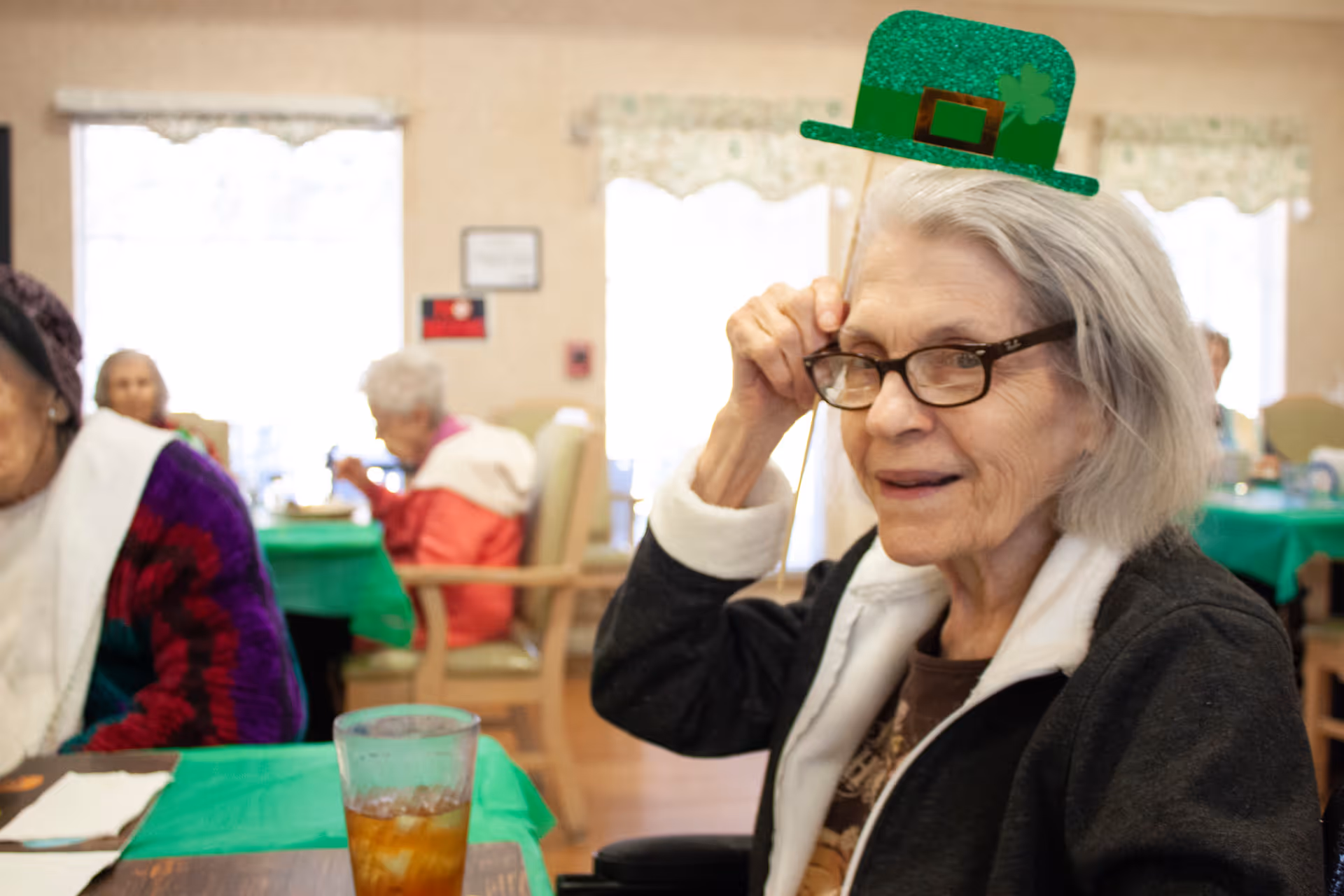 An elderly woman wearing glasses and a black jacket is sitting at a table in a dining area decorated with green tablecloths. She is holding a green glittery paper hat on a stick above her head. Other elderly individuals are seated at tables in the background.
