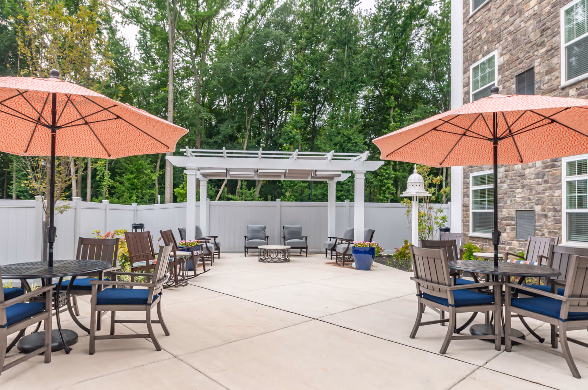 Outdoor patio with tables, orange umbrellas, chairs, a pergola, and potted plants beside a stone building.