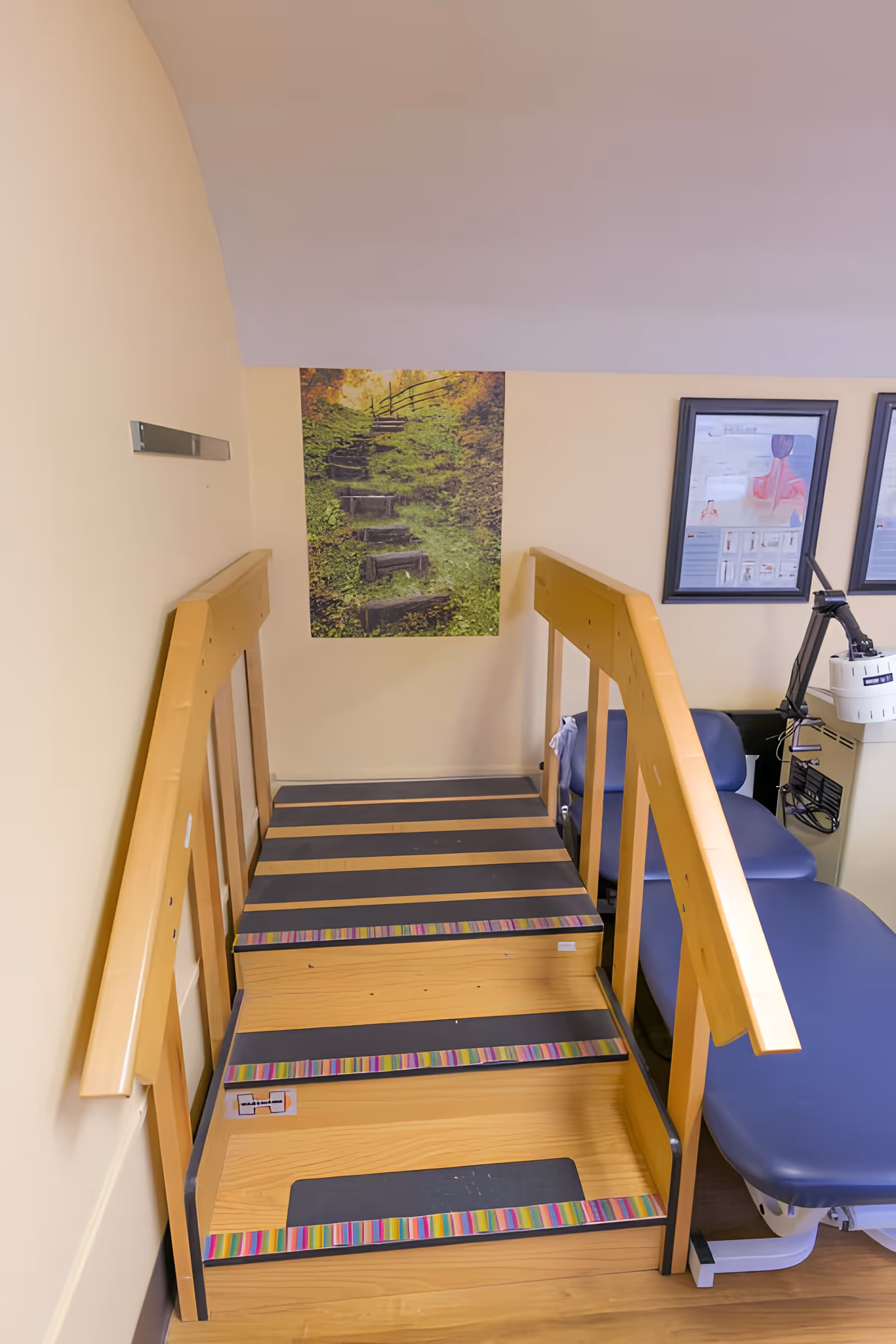 Wooden therapy stairs with handrails in a rehab room beside a treatment table and framed wall art.