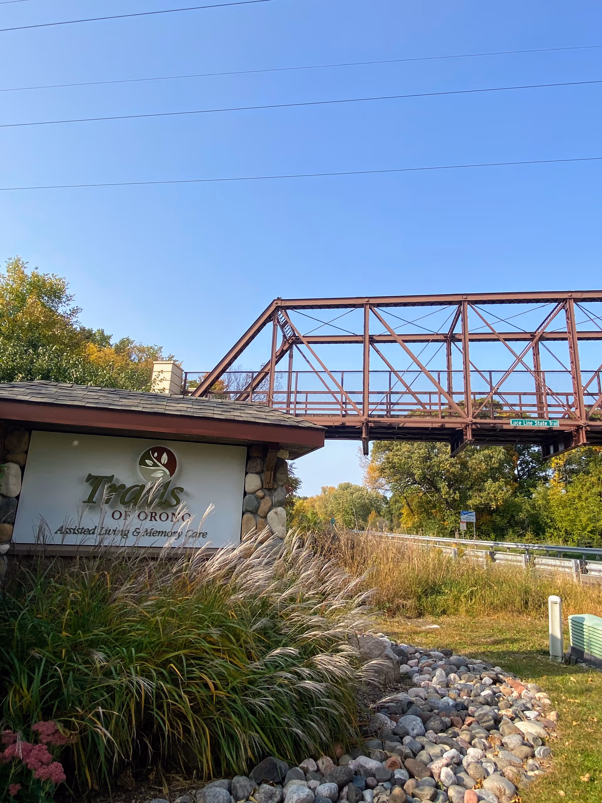 Outdoor view of the Trails of Orono Assisted Living & Memory Care sign surrounded by tall grass and rocks, with a rusty metal bridge and trees in the background under a clear blue sky.