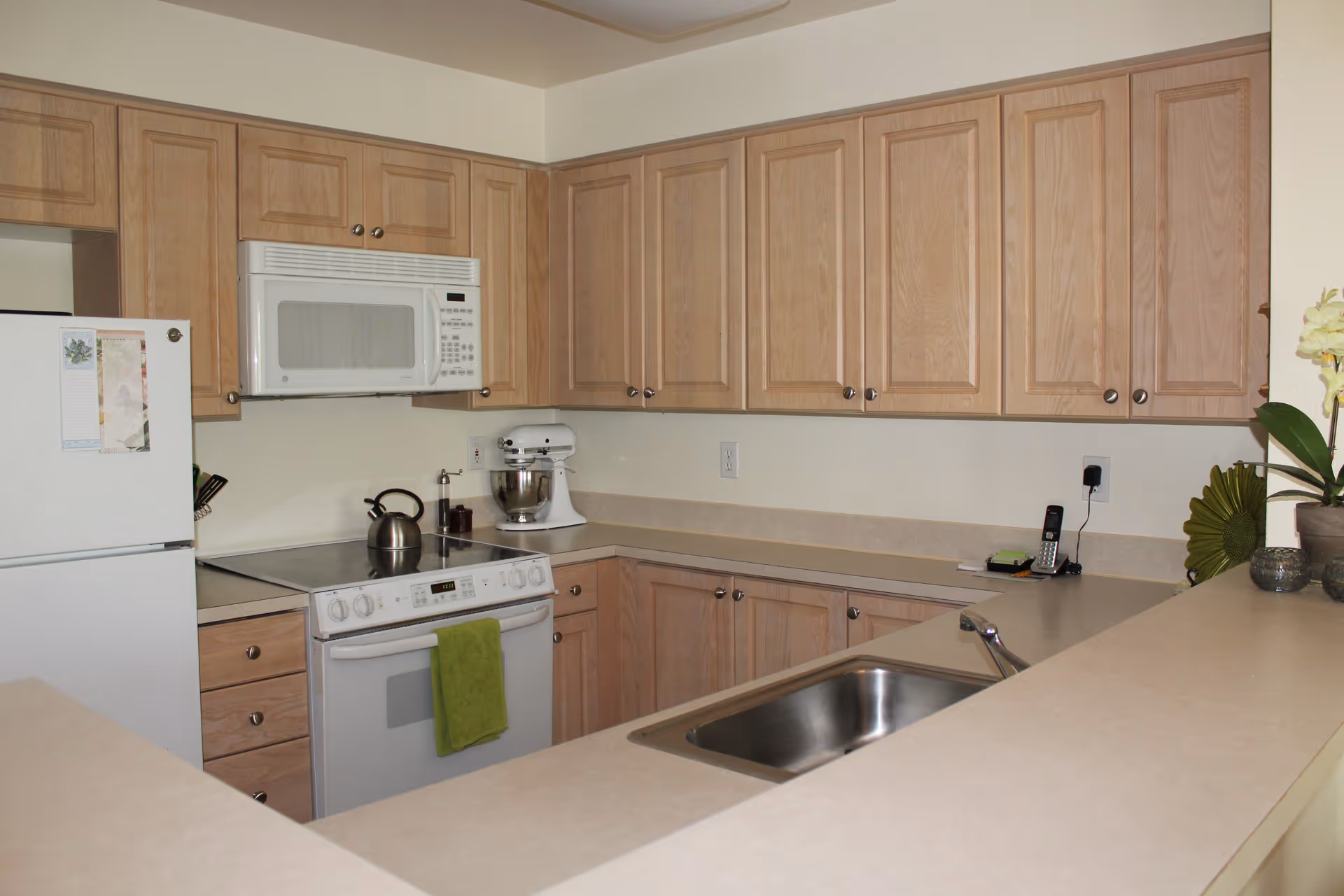 A kitchen with light wood cabinets, a white refrigerator, a white microwave mounted above a white stove with a green towel hanging on the oven handle, a stainless steel sink, and a countertop with a cordless phone and decorative plants.