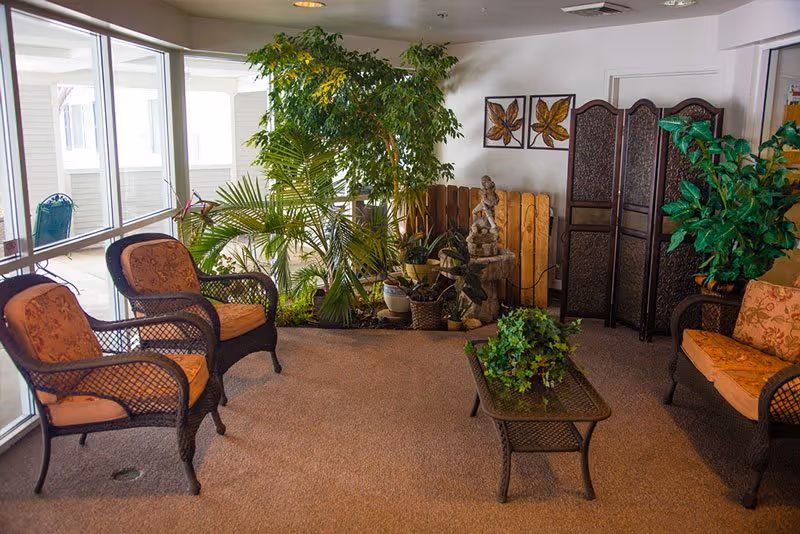 A cozy indoor seating area with wicker chairs and a loveseat featuring orange cushions, a glass-top coffee table with a green plant, several potted plants, a decorative wooden screen, and framed leaf artwork on the wall. Large windows allow natural light to fill the space.