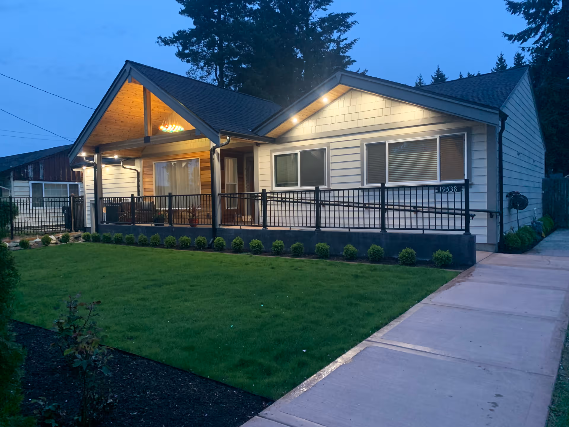 Exterior view of a single-story residential building at dusk with lights on under the porch roof and along the front. The building has white siding, a black metal railing along the porch, a well-maintained green lawn, and a concrete walkway leading to the entrance. Trees are visible in the background.