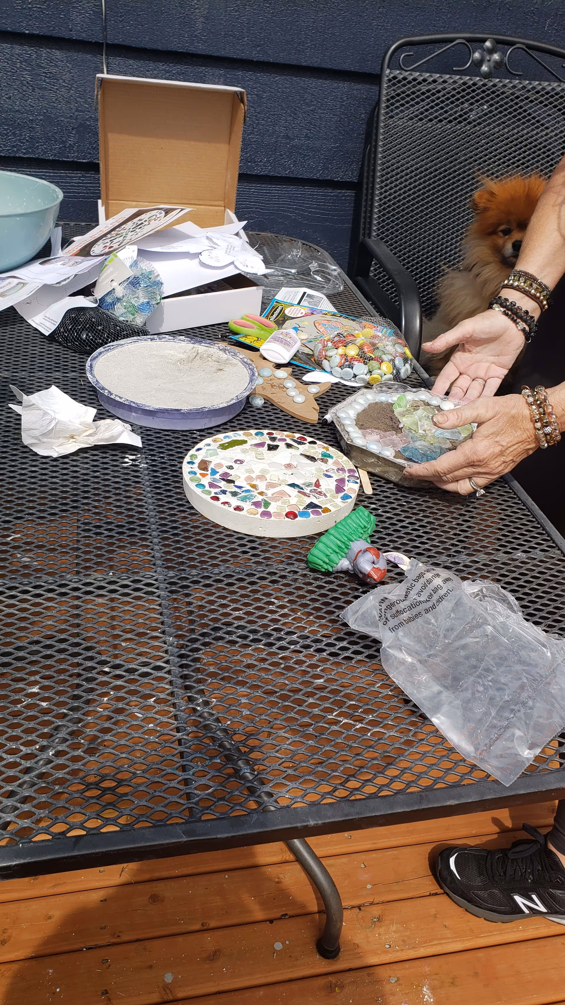 A metal outdoor table with craft supplies including mosaic pieces, glue, and bowls with cement or plaster. A person wearing bracelets is holding a mosaic craft piece. A small dog is partially visible sitting on a metal chair behind the person. The setting appears to be an outdoor deck with wooden flooring and a dark blue wall in the background.