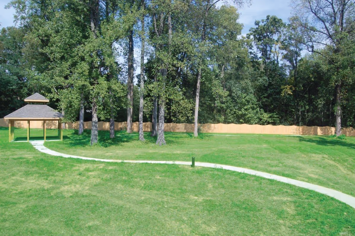 A green outdoor area with a curved concrete pathway leading to a small wooden gazebo. Tall trees and a wooden fence are visible in the background under a clear blue sky.