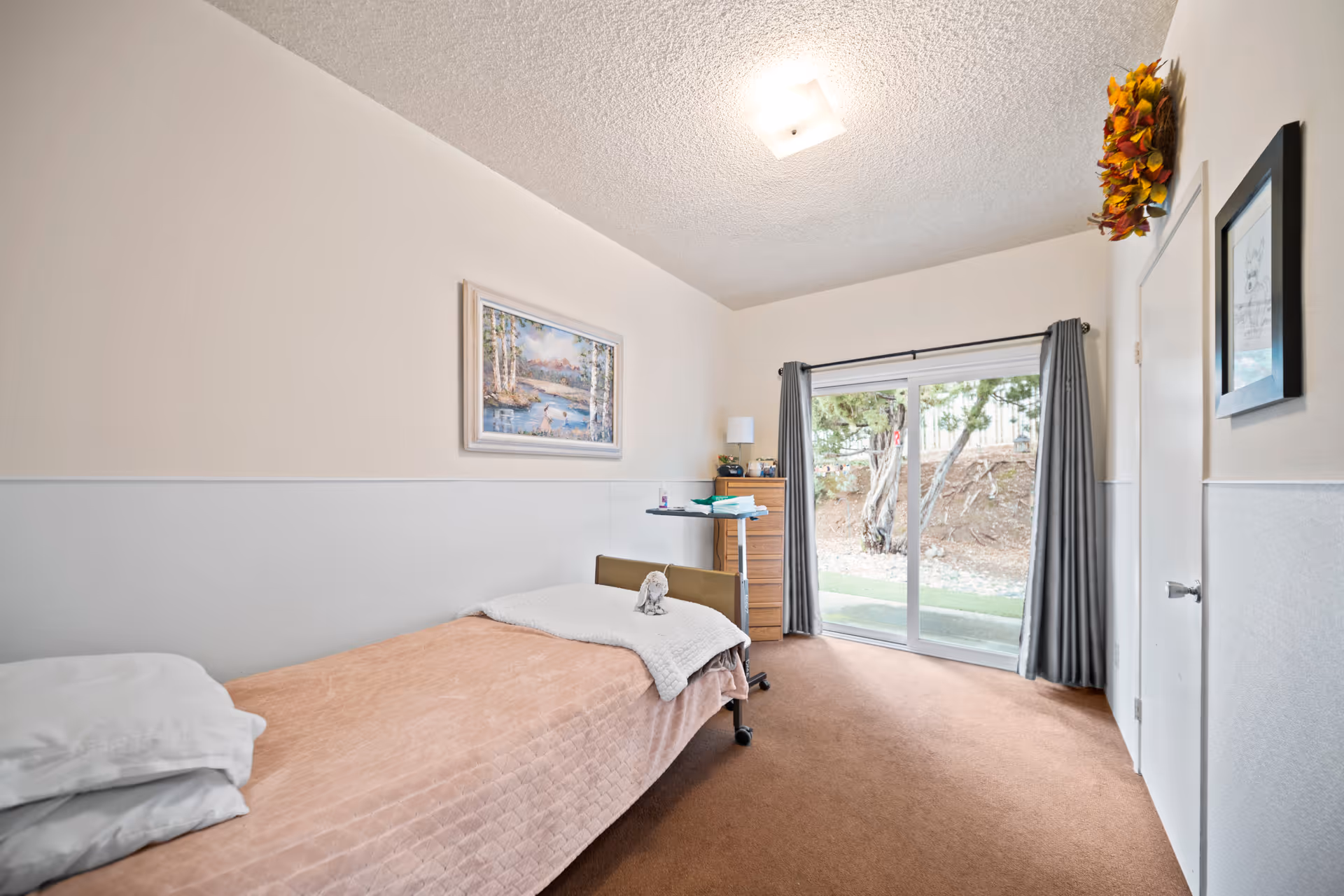 A simple bedroom with a single bed covered with a peach-colored blanket and white pillows. There is a small bedside table with a lamp and some items on it, a framed landscape painting on the wall, and a sliding glass door with gray curtains leading outside. The room has beige walls with white wainscoting and brown carpeted floor.