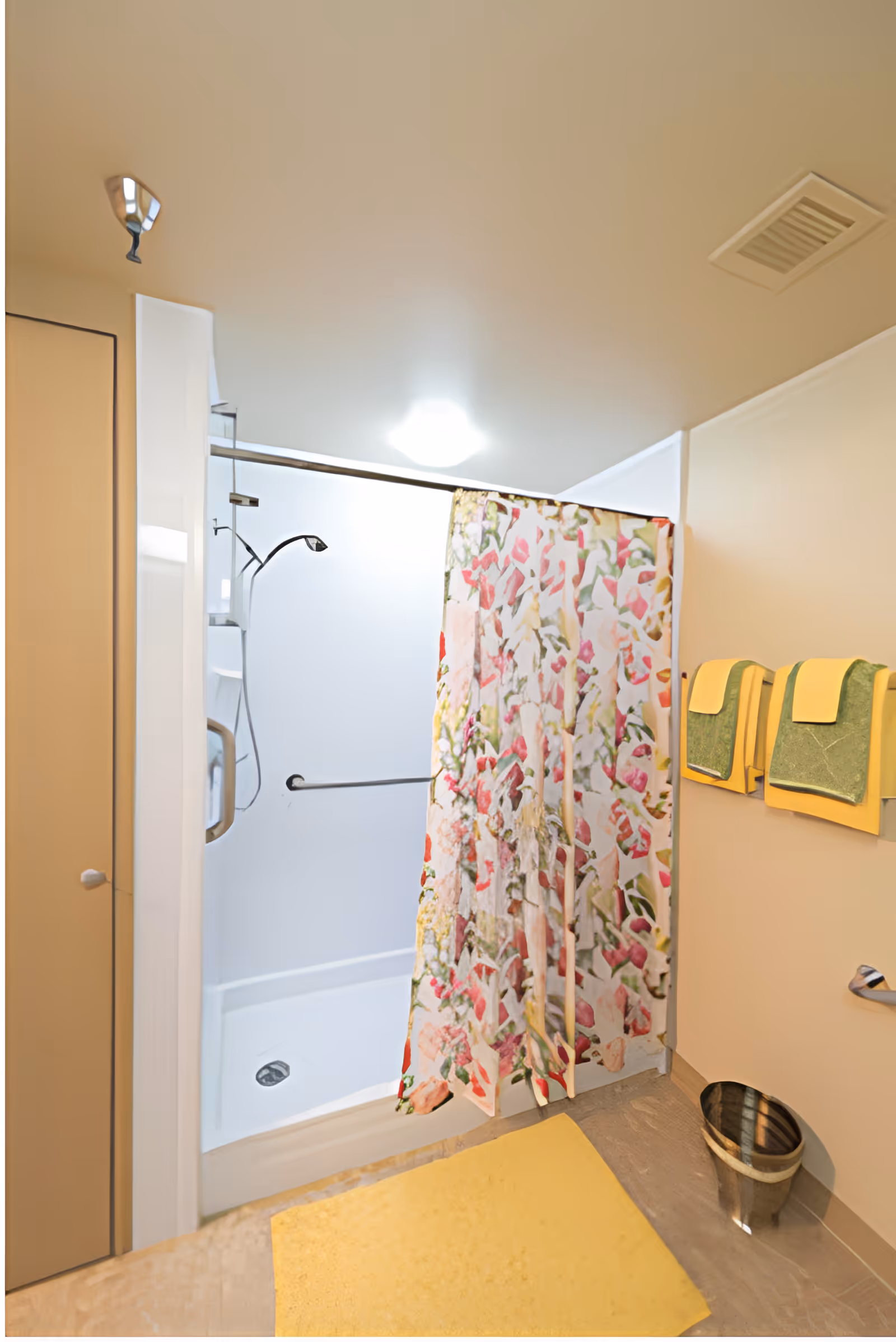 A bathroom shower area with a floral patterned shower curtain partially drawn. The shower has a white base and walls, a handheld showerhead, and a grab bar. On the wall outside the shower, there are two green and yellow towels hanging on towel racks. A yellow bath mat is on the floor, and a small metal trash can is placed nearby.