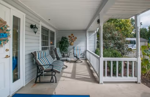 Covered outdoor porch area with striped cushioned chairs and a small table. The porch has white railings and columns, with a view of greenery and a parked vehicle in the background.
