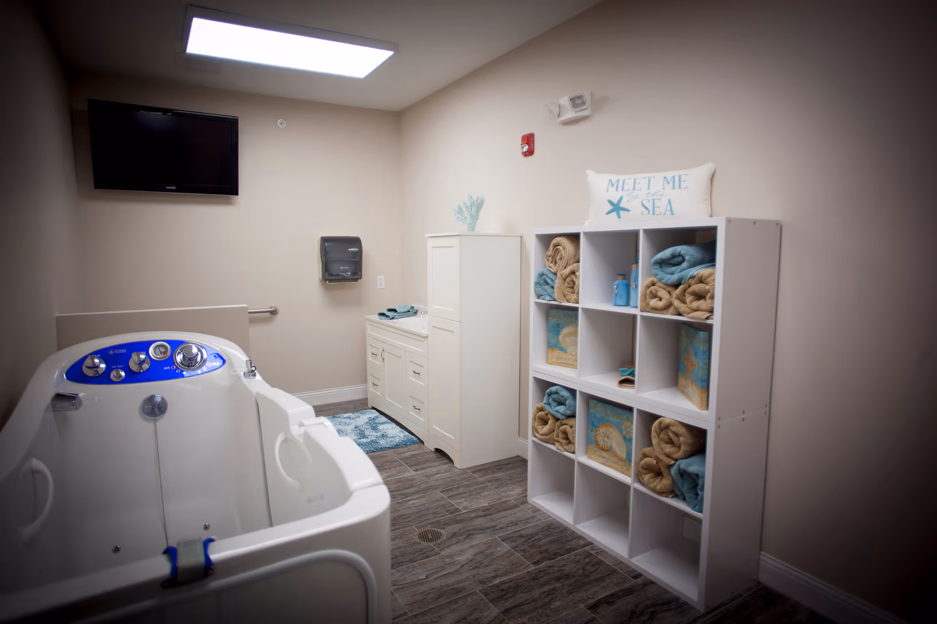A clean and well-lit assisted living bathing room featuring a white walk-in bathtub with blue control panel, a white storage cabinet with towels and decorative items, a wall-mounted TV, and a paper towel dispenser. The room has beige walls and wood-look tile flooring.