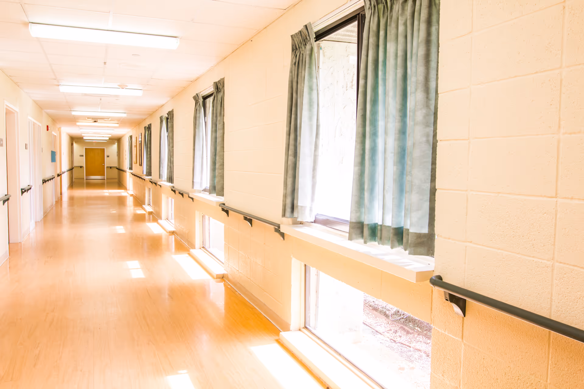 A brightly lit hallway in a nursing and rehabilitation center with light-colored walls, wooden flooring, handrails along both sides, and windows with green curtains letting in natural light.