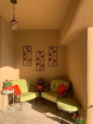 A cozy indoor seating area with two light green cushioned chairs, a matching cushioned ottoman, and a small round table with a potted plant. The walls are beige, decorated with three vertical floral wall hangings. A hanging light fixture is visible on the ceiling.