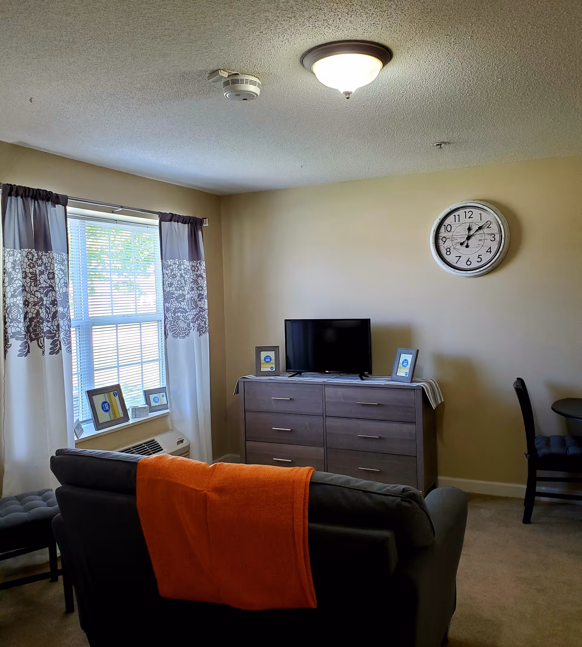 A cozy living room with a gray couch draped with an orange blanket facing a wooden dresser with a flat-screen TV on top. There are three framed pictures on the dresser. A large window with white and gray patterned curtains lets in natural light. A round wall clock hangs on the beige wall above the dresser. A small dining table with two chairs is partially visible on the right side.