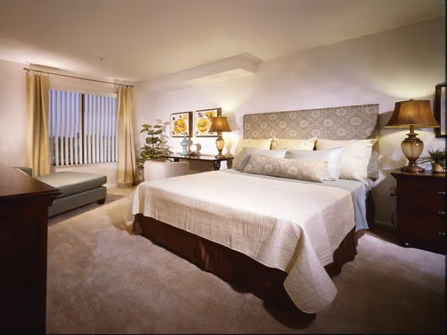 A well-lit bedroom with a large bed, patterned headboard, matching nightstands and lamps, a chaise by a window, and framed artwork on the wall.