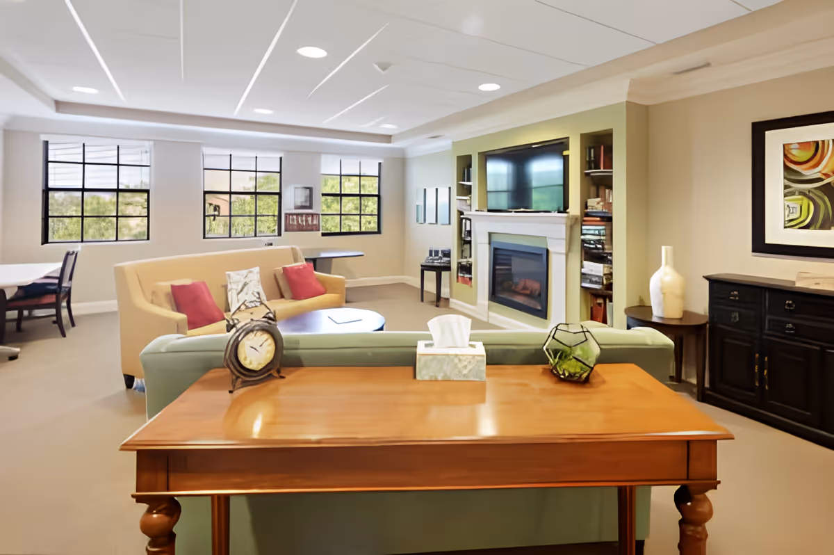 A cozy living room area with a wooden table in the foreground holding a clock, tissue box, and decorative item. Behind the table is a green sofa facing a beige couch with red pillows. The room features large windows letting in natural light, a fireplace with a TV above it, built-in shelves, and a dark wooden cabinet with a vase and framed artwork on the wall.