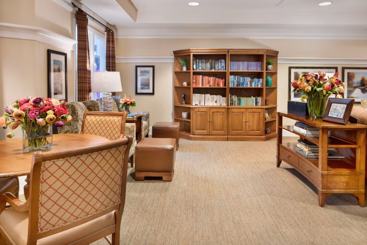 Bright common room with wooden bookshelves, upholstered chairs, a dining table, and floral arrangements.