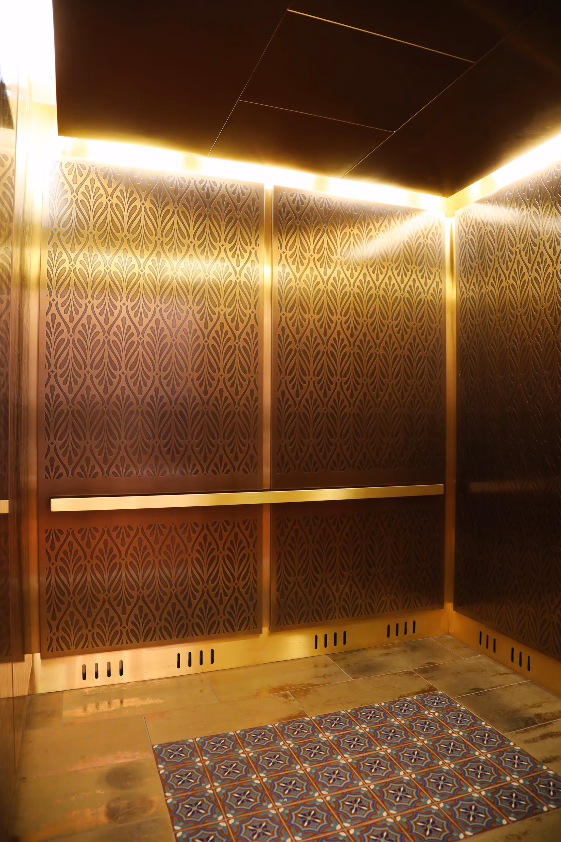 Interior of an elevator with ornate patterned bronze walls, a horizontal handrail, and a section of decorative tiled floor in front.
