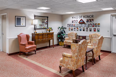 A sitting area in a senior living facility with four patterned armchairs arranged around a small wooden table on a red and beige carpet. Against the wall is a wooden console table with a lamp, flowers, and a mirror above it. On the wall to the right is a 'Wall of Honor' display featuring framed photos and a folded American flag. There is a potted plant in the corner and two closed doors on either side of the room.