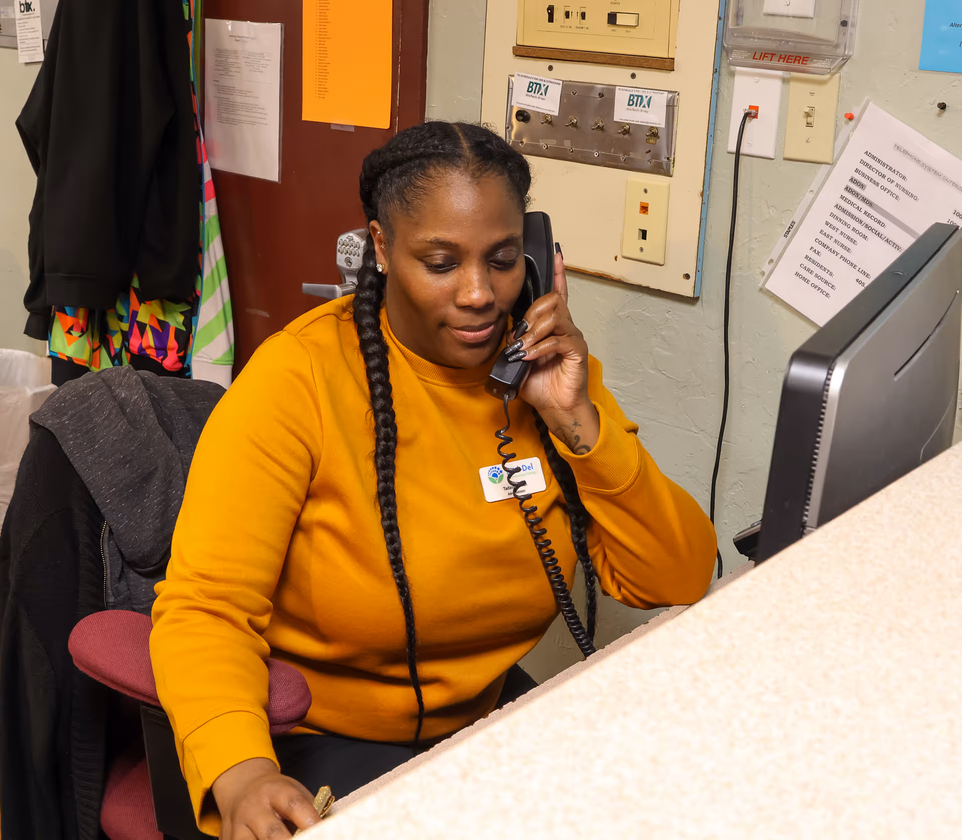 A woman with long braided hair wearing a mustard yellow sweater is sitting at a desk, talking on a corded phone while using a computer. Behind her are various papers, a coat hanging on a chair, and a wall with electrical panels and notices.