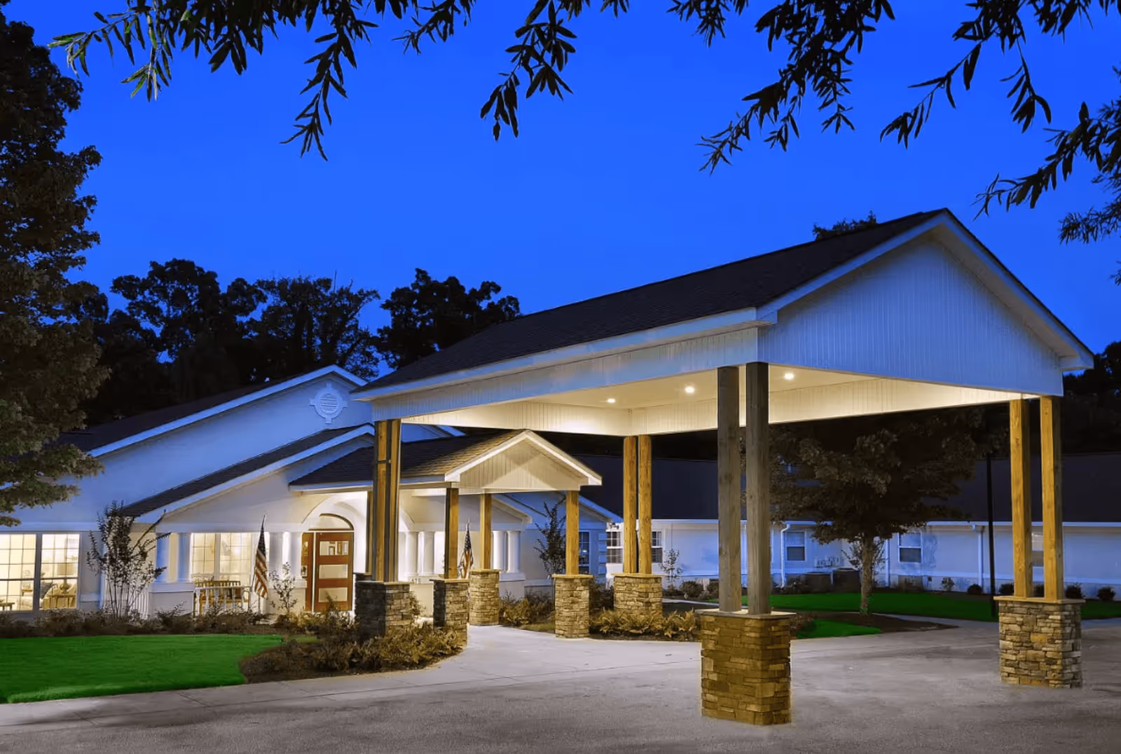 Exterior view of The Waterford at Decatur senior living facility at dusk, showing a covered entrance with stone pillars and a well-lit building surrounded by trees and landscaping.