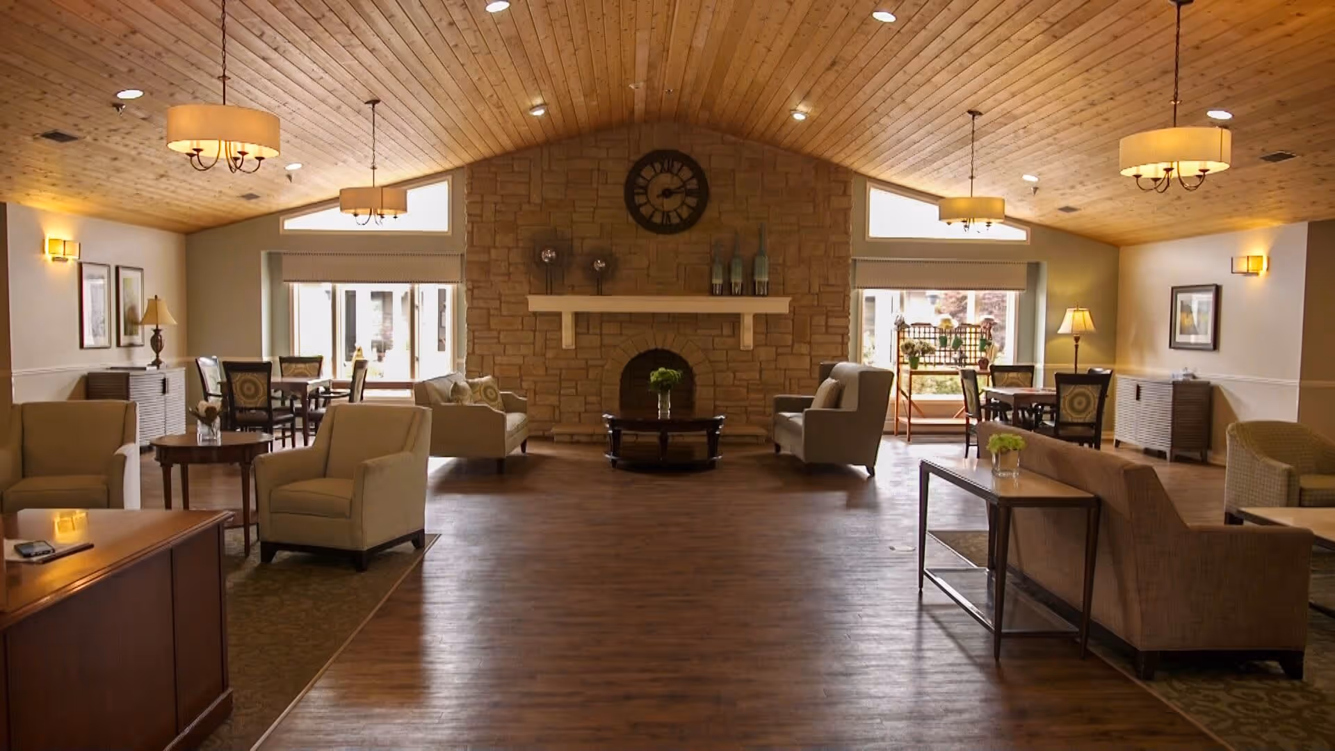 A spacious living room area with a high wooden ceiling and wooden floor. The room features a stone fireplace with a clock above it, surrounded by comfortable armchairs and sofas. There are several tables with chairs near large windows letting in natural light. The room is warmly lit with ceiling lights and wall sconces, decorated with framed pictures and small plants on tables.