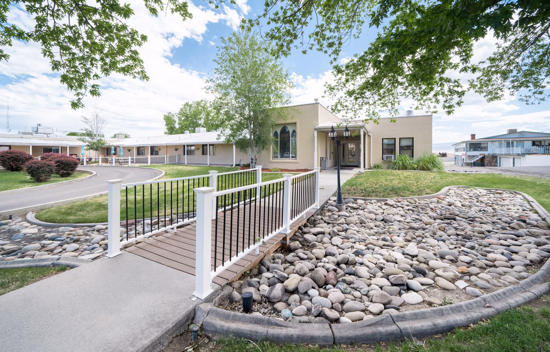 Exterior view of Mantey Heights Rehabilitation and Care Center showing a single-story building with a beige facade, a small bridge over a dry rock bed, green grass, trees, and a partly cloudy sky.
