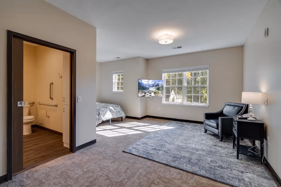 A bright senior living room with a large window letting in natural light. The room features a black armchair next to a small black side table with a lamp. A flat-screen TV is mounted on the wall opposite the window. To the left, there is an open doorway leading to a bathroom with grab bars and a toilet visible. The room has carpeted flooring and neutral-colored walls.