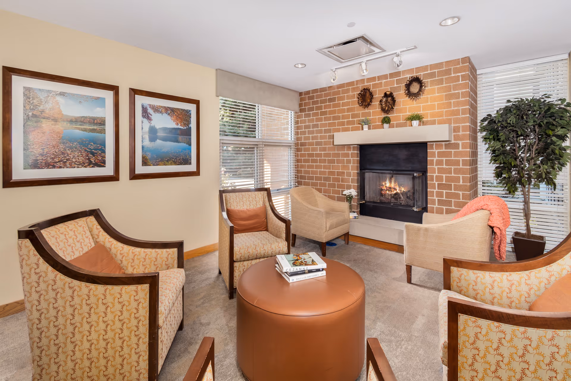 A cozy living room with a brick fireplace and a fire burning inside. The room has four upholstered armchairs arranged around a round brown ottoman with books on top. There are two framed landscape photos on the wall, large windows with blinds, and a tall potted plant near the fireplace. The room is warmly lit and inviting.