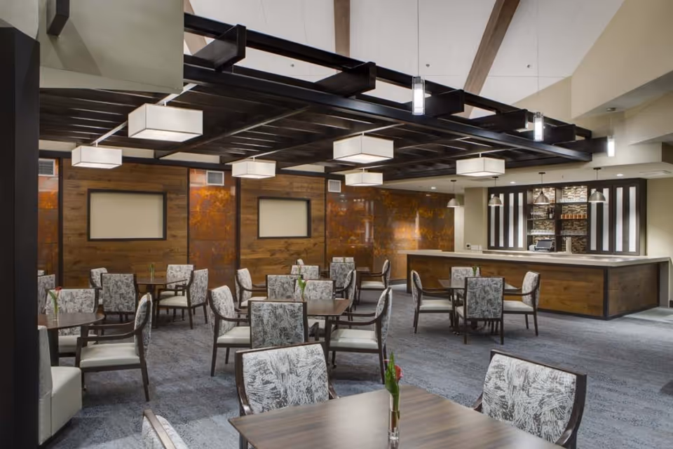 A modern dining room with multiple square wooden tables and cushioned chairs featuring a gray and white abstract pattern. The room has a high ceiling with exposed beams and contemporary rectangular pendant lights. There is a wooden bar area with a stone backsplash and pendant lights in the background. The walls are decorated with wood paneling and rust-colored accents.