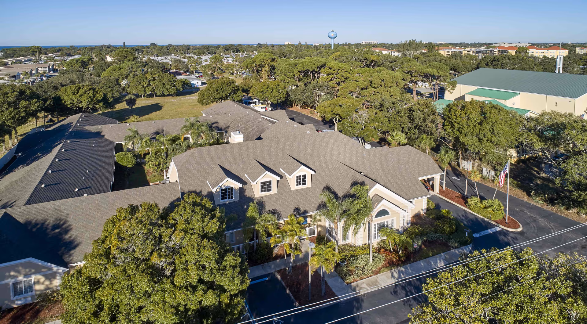 Aerial view of Maris Pointe senior living facility surrounded by trees and greenery, showing a large building with a gray roof, multiple windows, and a landscaped entrance with palm trees and an American flag.