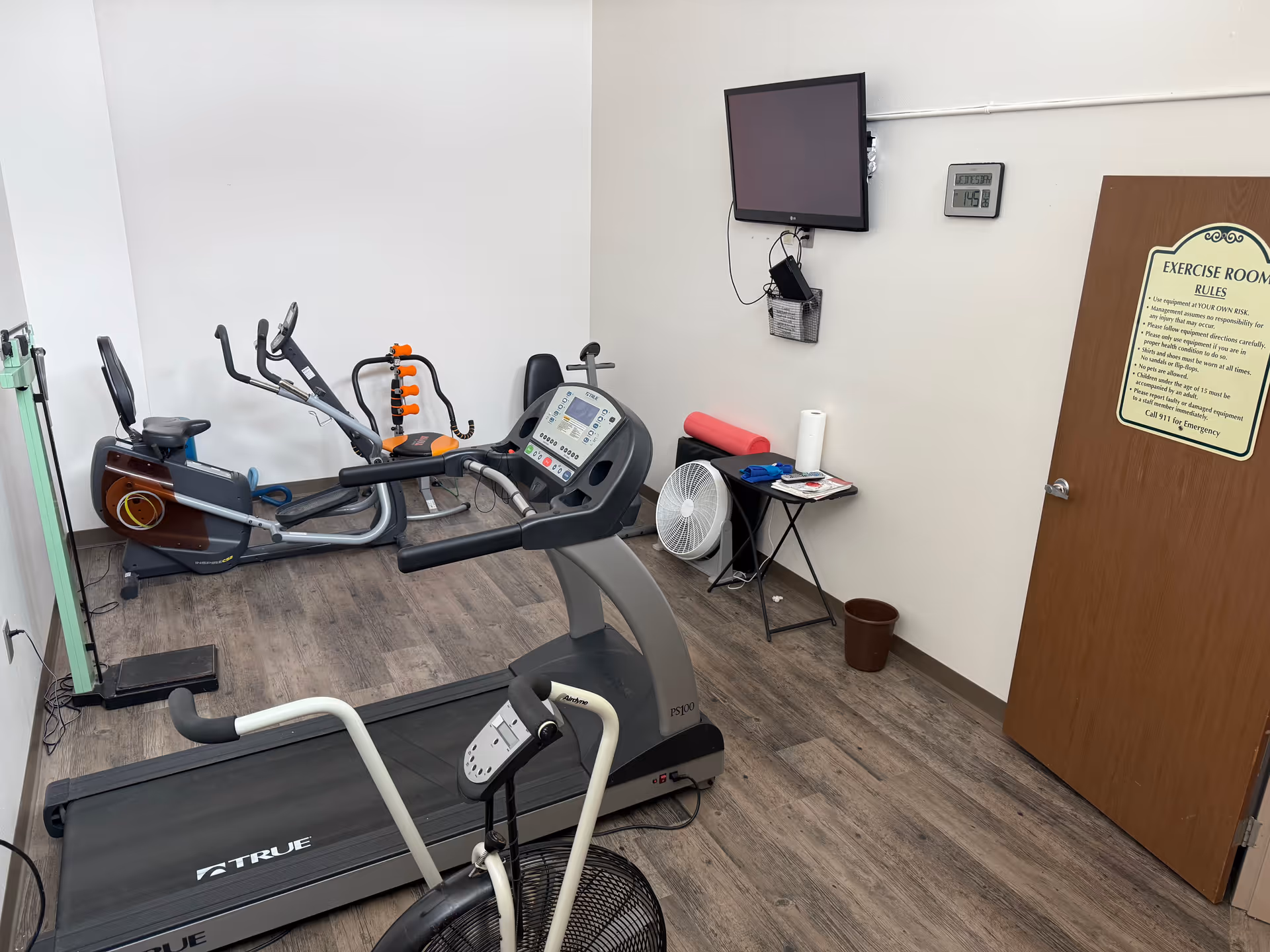 Small exercise room with a treadmill, stationary bike, elliptical machine, and a rack of dumbbells. A wall-mounted TV and a clock are visible above a small table holding a fan, paper towel roll, and some exercise accessories. A door with exercise room rules is on the right side.