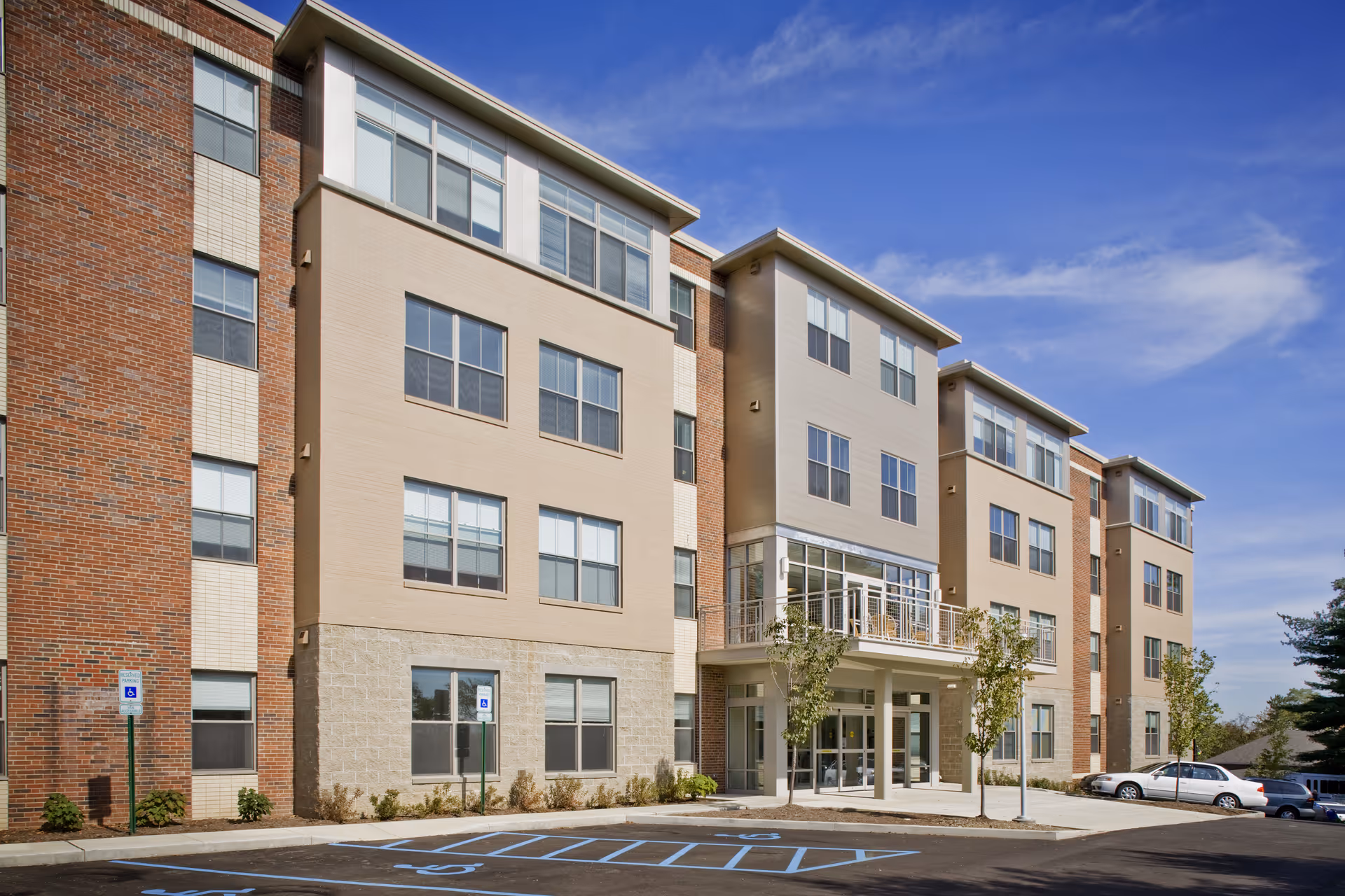 Exterior view of a modern multi-story retirement residence building with large windows, a main entrance with glass doors, small trees planted near the entrance, and a parking lot with handicap parking spaces in front under a partly cloudy blue sky.