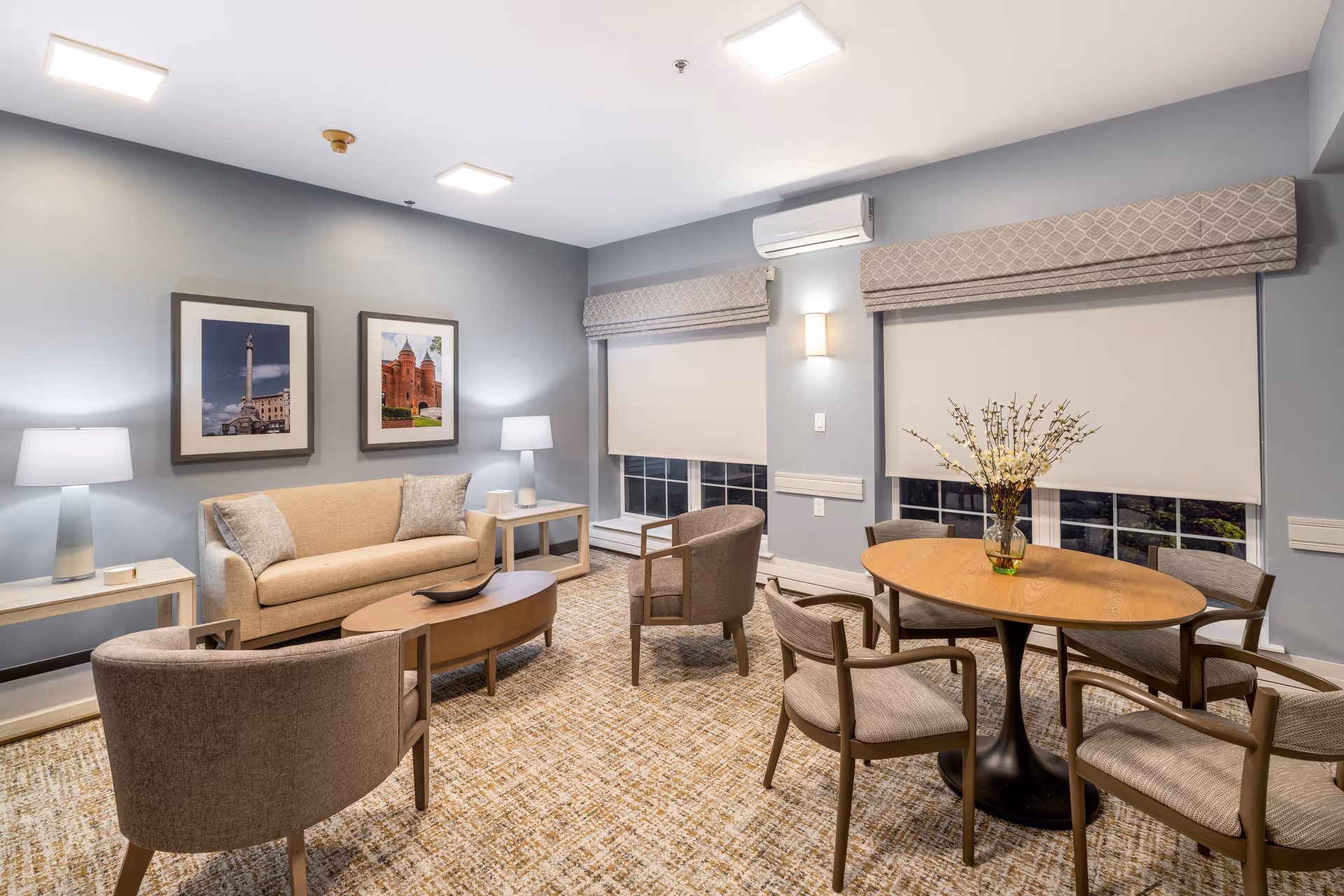 A cozy senior living community room with a beige sofa, two armchairs, a wooden coffee table, and a round dining table with four chairs. The walls are painted light gray and decorated with two framed pictures. The room has large windows with beige roller shades and patterned valances, soft lighting from table lamps and wall sconces, and a patterned carpet.