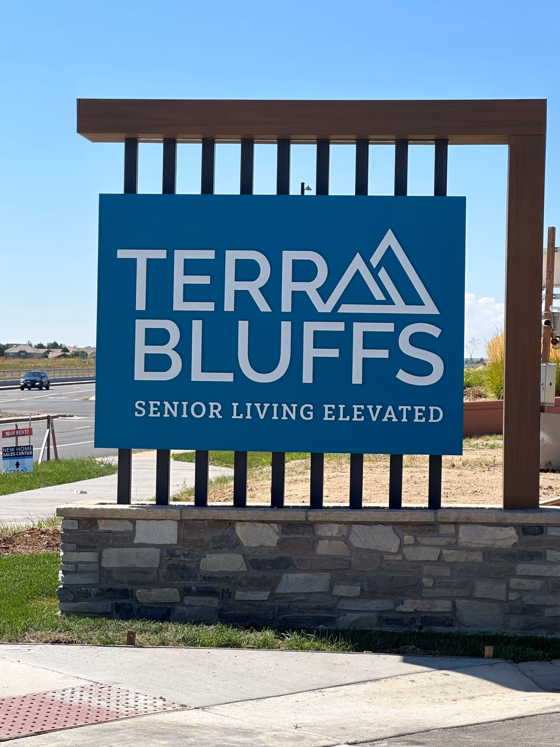 Outdoor sign for Terra Bluffs senior living community with the tagline 'Senior Living Elevated' displayed on a blue background, mounted on a stone base near a sidewalk and road.