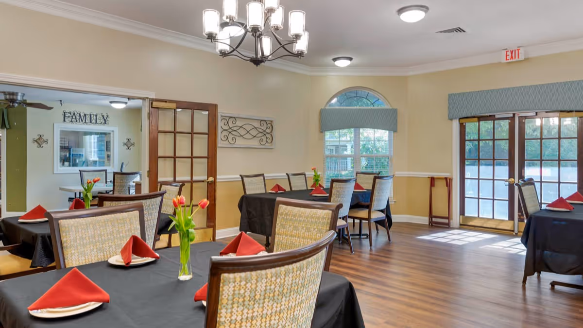 A bright dining room with several tables covered in black tablecloths, each set with white plates and red folded napkins. Small vases with red tulips decorate the tables. The room has wooden floors, beige walls, large windows with blue valances, and a chandelier hanging from the ceiling. French doors lead outside, and a wall decoration with the word 'FAMILY' is visible in an adjacent room.