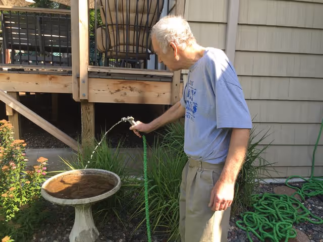 An elderly man wearing a gray t-shirt and beige pants waters a birdbath with a green garden hose in a backyard garden area next to a wooden deck and house siding.