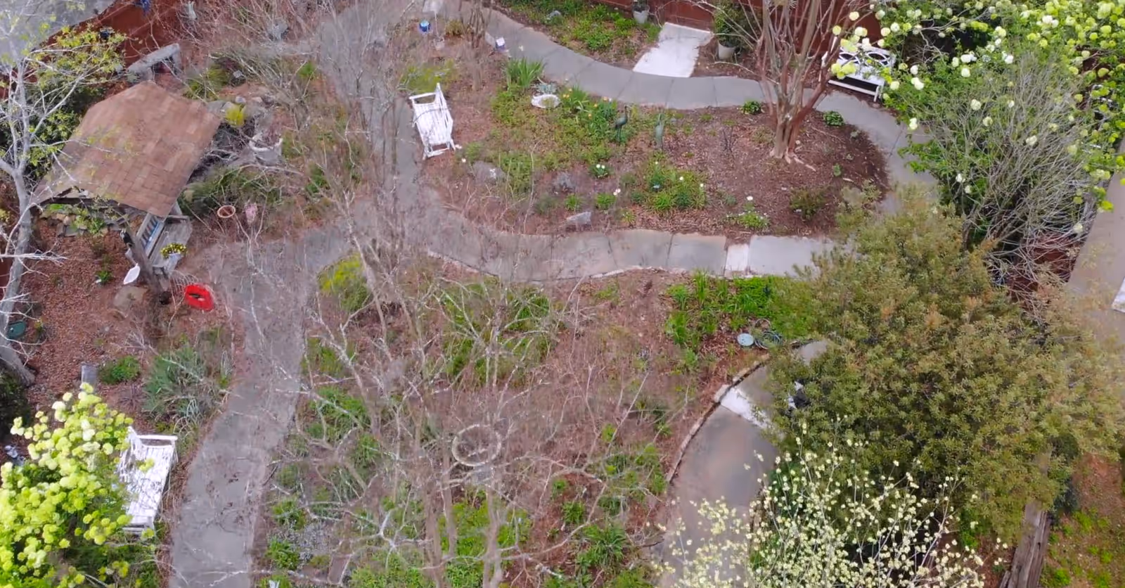 Aerial view of a garden area with winding concrete pathways, leafless trees, green shrubs, and white benches. There is a small wooden structure with a tiled roof on the left side and various plants and flowers scattered throughout the garden.