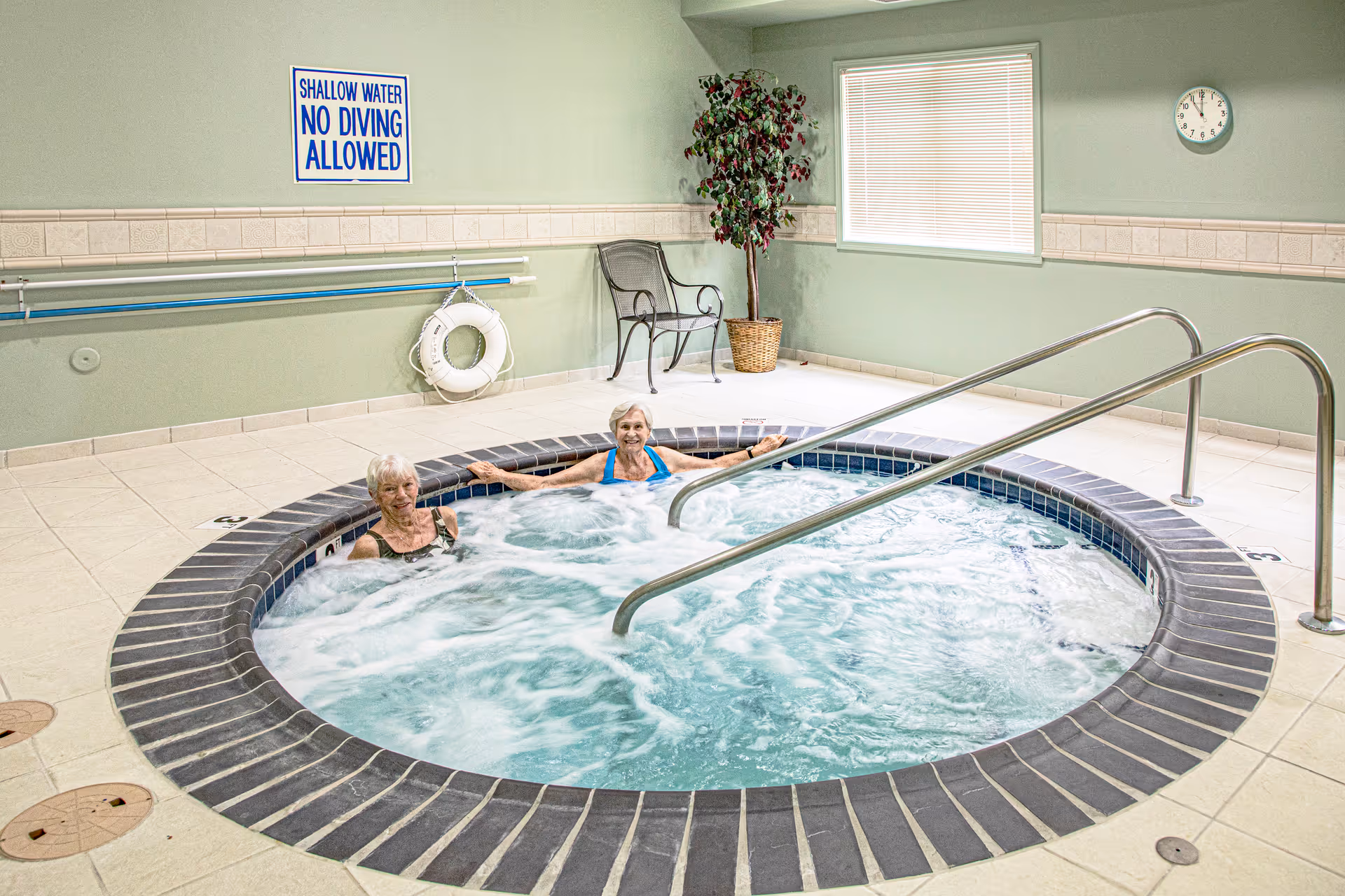 Two elderly women relaxing in a circular indoor hot tub with bubbling water. The room has light green walls, a clock on the wall, a potted plant, a chair, and a sign that reads 'SHALLOW WATER NO DIVING ALLOWED'.