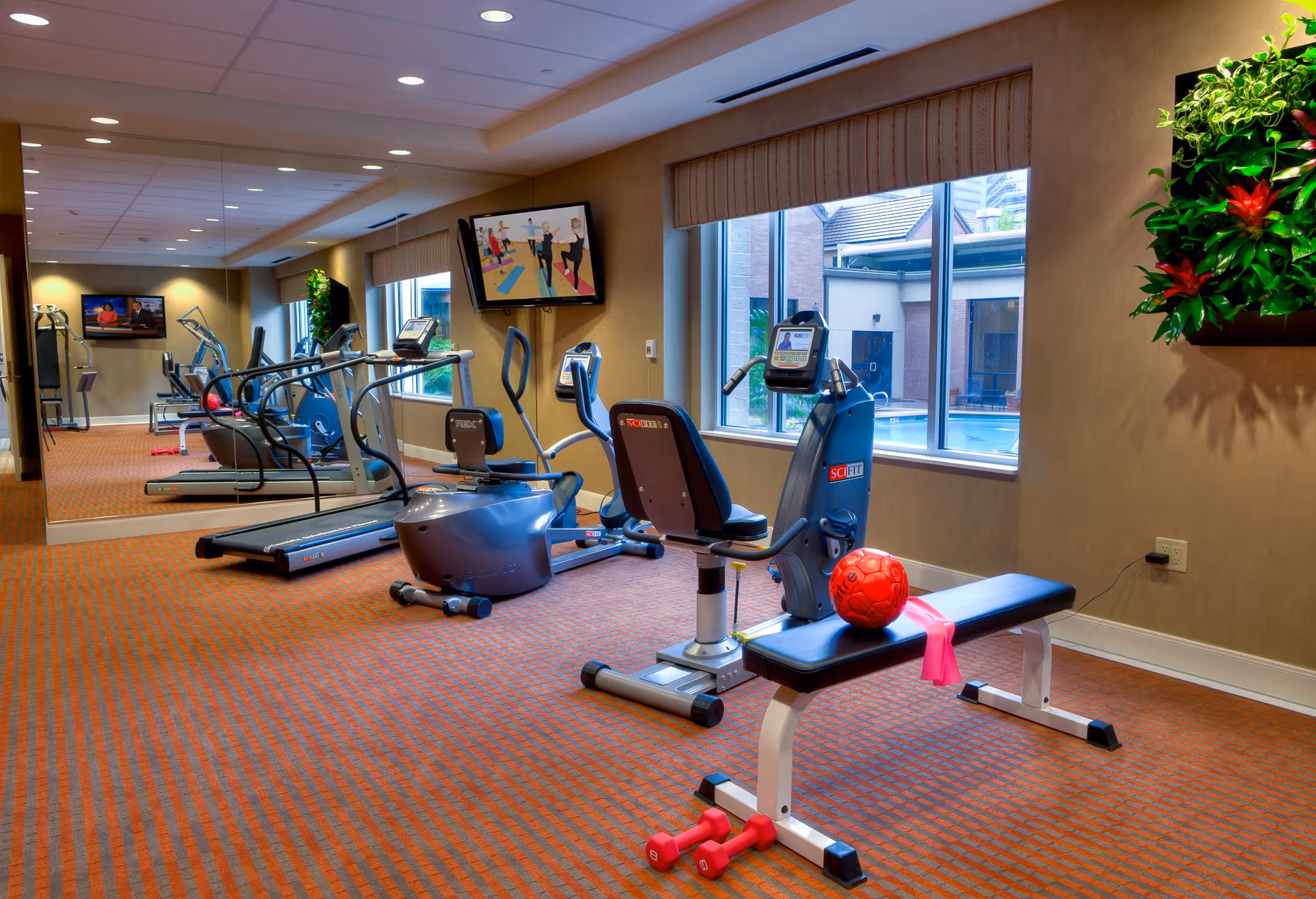 A fitness room with exercise equipment including treadmills, stationary bikes, and a bench with a red exercise ball and pink resistance band. The room has large windows showing an outdoor pool area, a wall-mounted TV, and a large mirror reflecting the equipment.