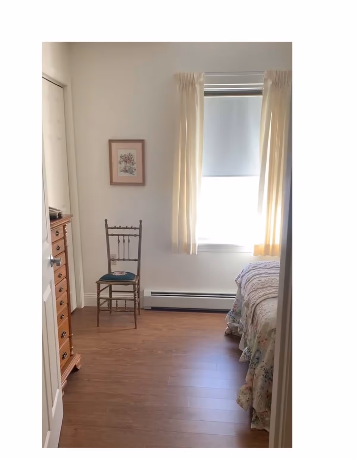 A small bedroom with wooden flooring, a wooden chair with a cushioned seat, a wooden dresser with multiple drawers, a bed with a floral bedspread partially visible on the right, a window with cream-colored curtains and a roller shade, and a framed picture hanging on the wall.