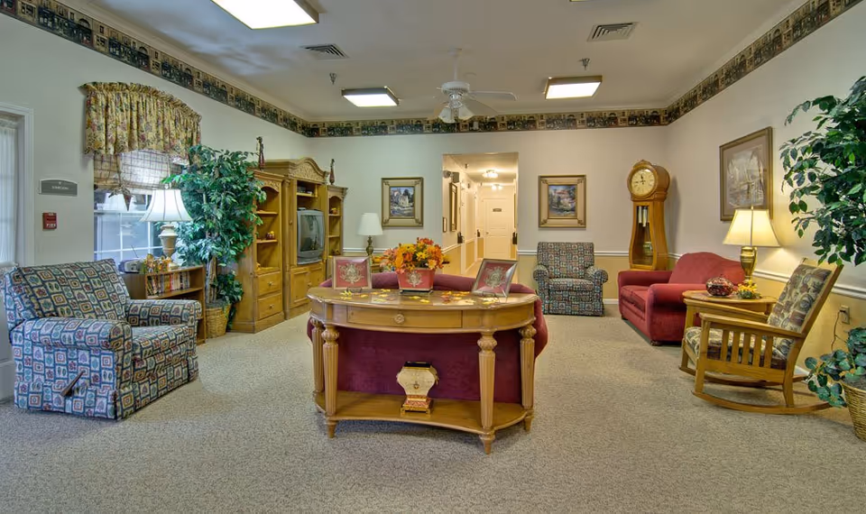 A cozy senior living facility common area with patterned armchairs, a red sofa, wooden rocking chair, wooden entertainment center with a TV, a grandfather clock, framed paintings on the walls, potted plants, and a wooden table with decorative items in the center.