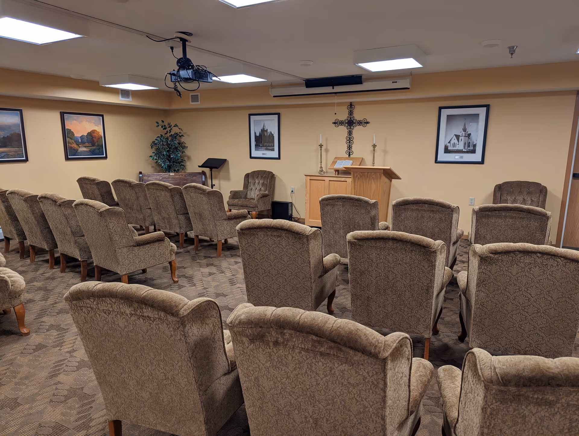 Rows of upholstered armchairs facing a small wooden podium with a decorative cross and framed artwork on the walls in a carpeted meeting/chapel room.