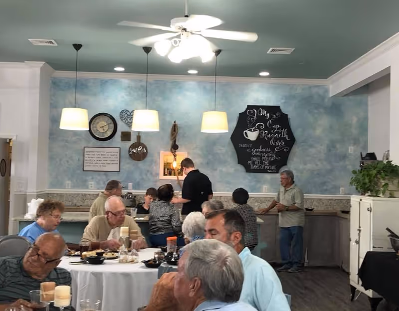 A group of elderly people sitting and standing around tables in a dining area with blue textured walls, pendant lights, a ceiling fan, and decorative wall art including a clock and a chalkboard sign with a coffee cup illustration.
