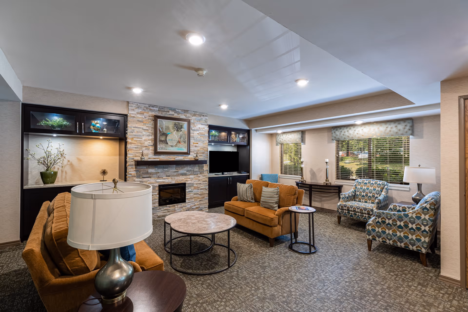 A cozy senior living community lounge with two brown sofas, two patterned armchairs, a round coffee table, side tables with lamps, a stone fireplace with a framed bicycle artwork above it, built-in dark wood cabinets with decorative items, and two windows with blinds letting in natural light.
