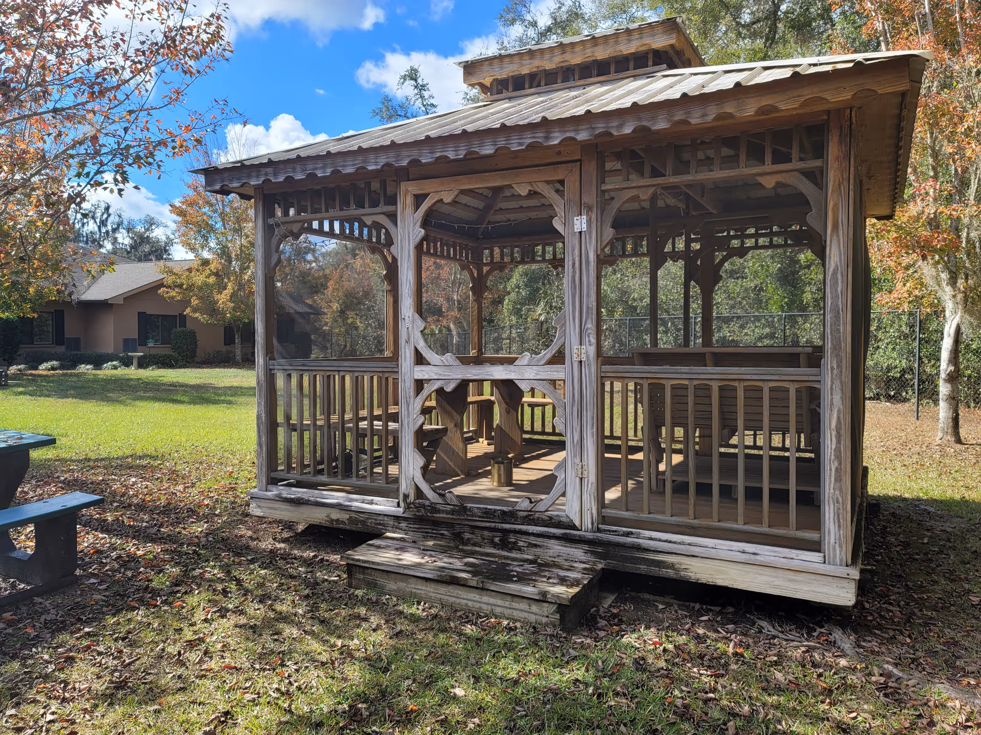 A wooden gazebo with a shingled roof situated outdoors on a grassy area with trees showing autumn foliage. There is a picnic table to the left and a building in the background under a partly cloudy blue sky.