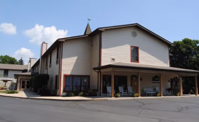 Exterior view of a two-story senior living facility building with beige siding and brown trim. The building has a covered porch area with several rocking chairs and potted plants. The sky is clear with a few clouds, and there are trees in the background.