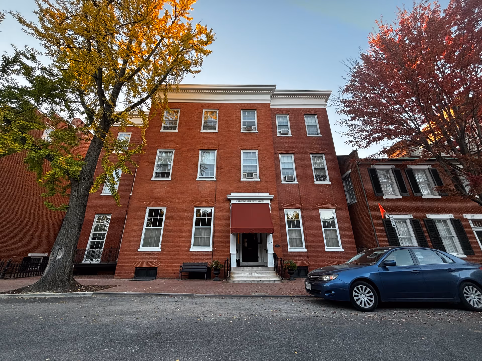 Front exterior view of a three-story red brick building with white window frames and a red awning above the entrance. There are two large trees with autumn-colored leaves on either side of the building and a blue car parked on the street in front.