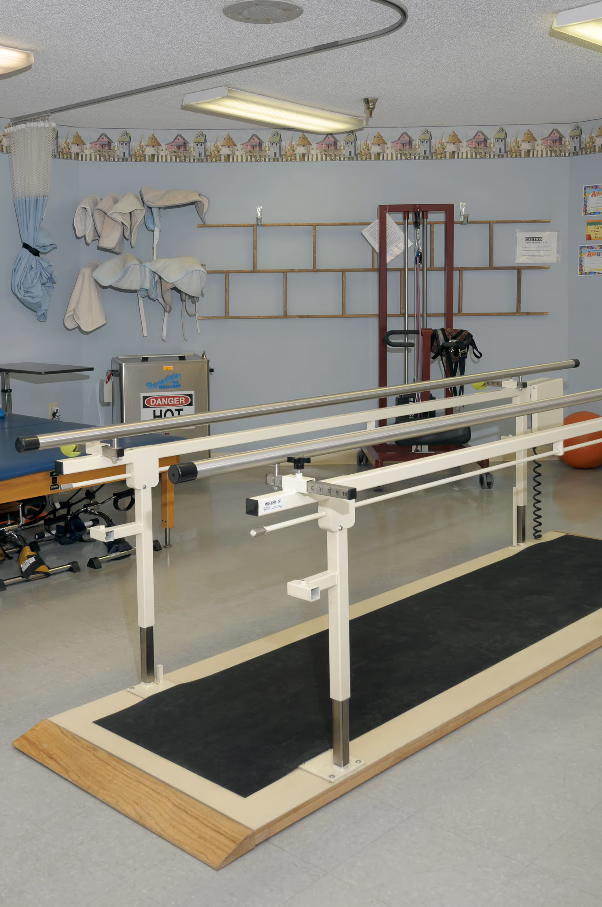 Rehabilitation therapy room featuring parallel walking bars on a platform, exercise equipment, and folded therapy towels on the wall.