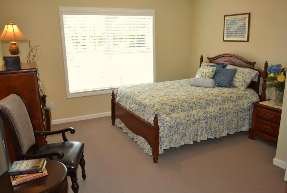 A cozy bedroom with a wooden bed frame and floral bedspread. The bed has several pillows and a bolster. There is a wooden nightstand with a vase of flowers and a framed picture on the wall above the bed. A window with white blinds lets in natural light. A wooden chair and a small round table with books are positioned near a wooden dresser with a lamp on top.
